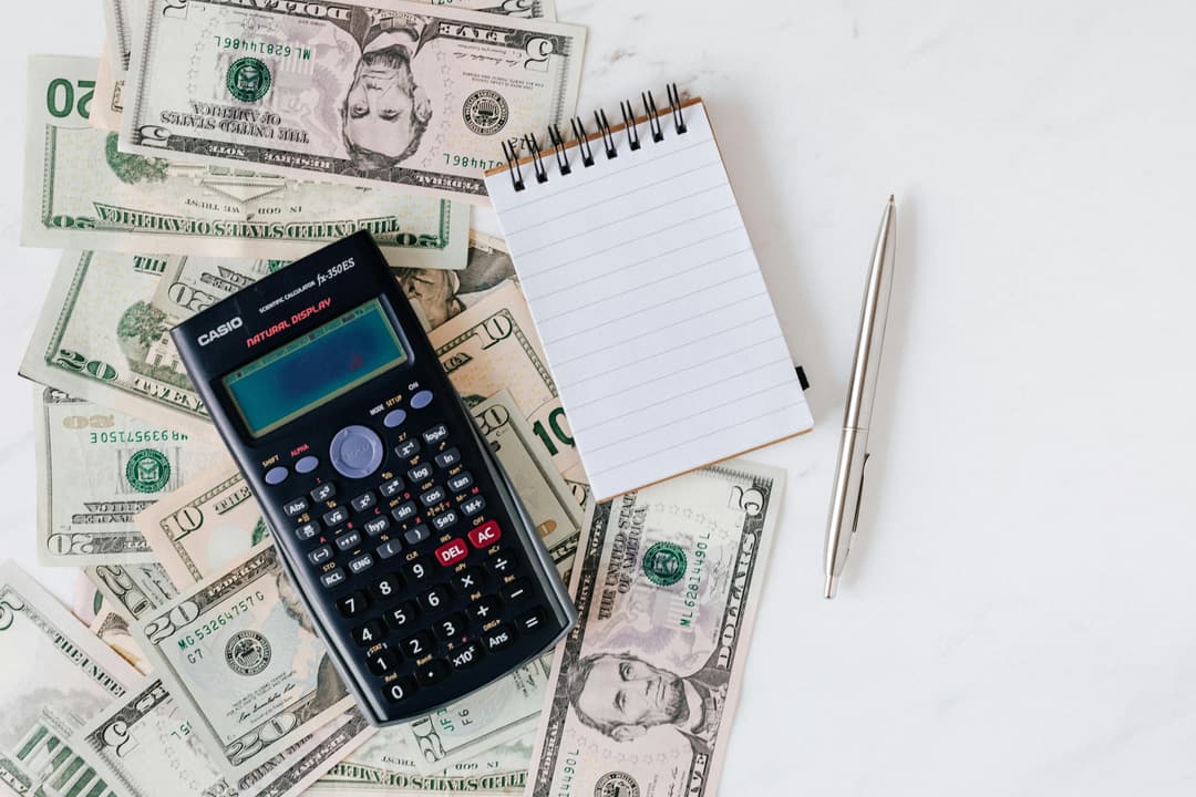 A calculater with paper and pen on top of a couple dollar bills